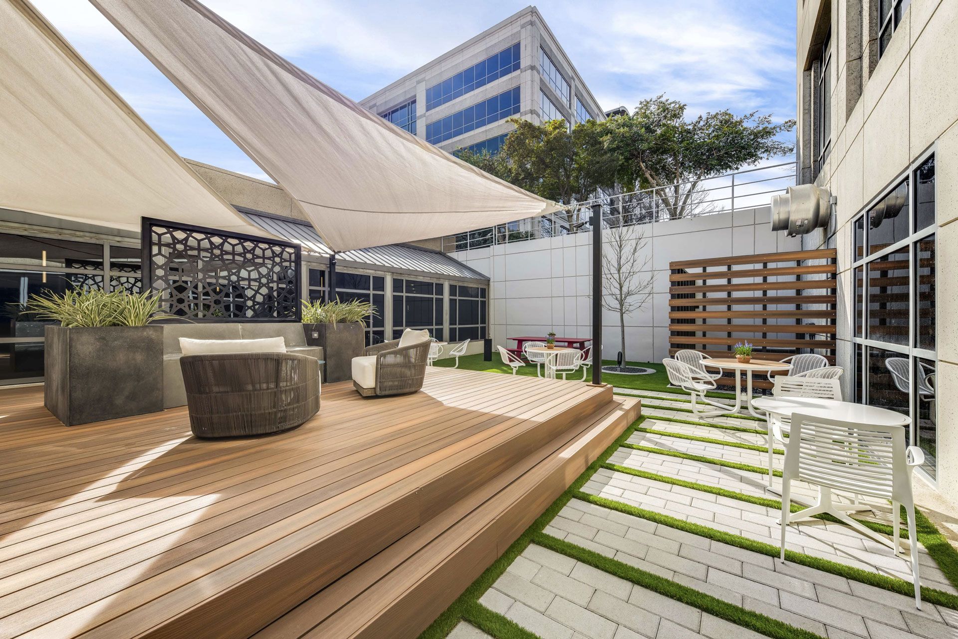 Outdoor patio at the Las Colinas Corporate Center, featuring a large canopy providing shade over seating areas. The space includes modern patio furniture, with tables and chairs arranged for relaxation or casual meetings. Surrounded by landscaped greenery and offering a view of the office building, the patio creates a comfortable and inviting atmosphere for tenants