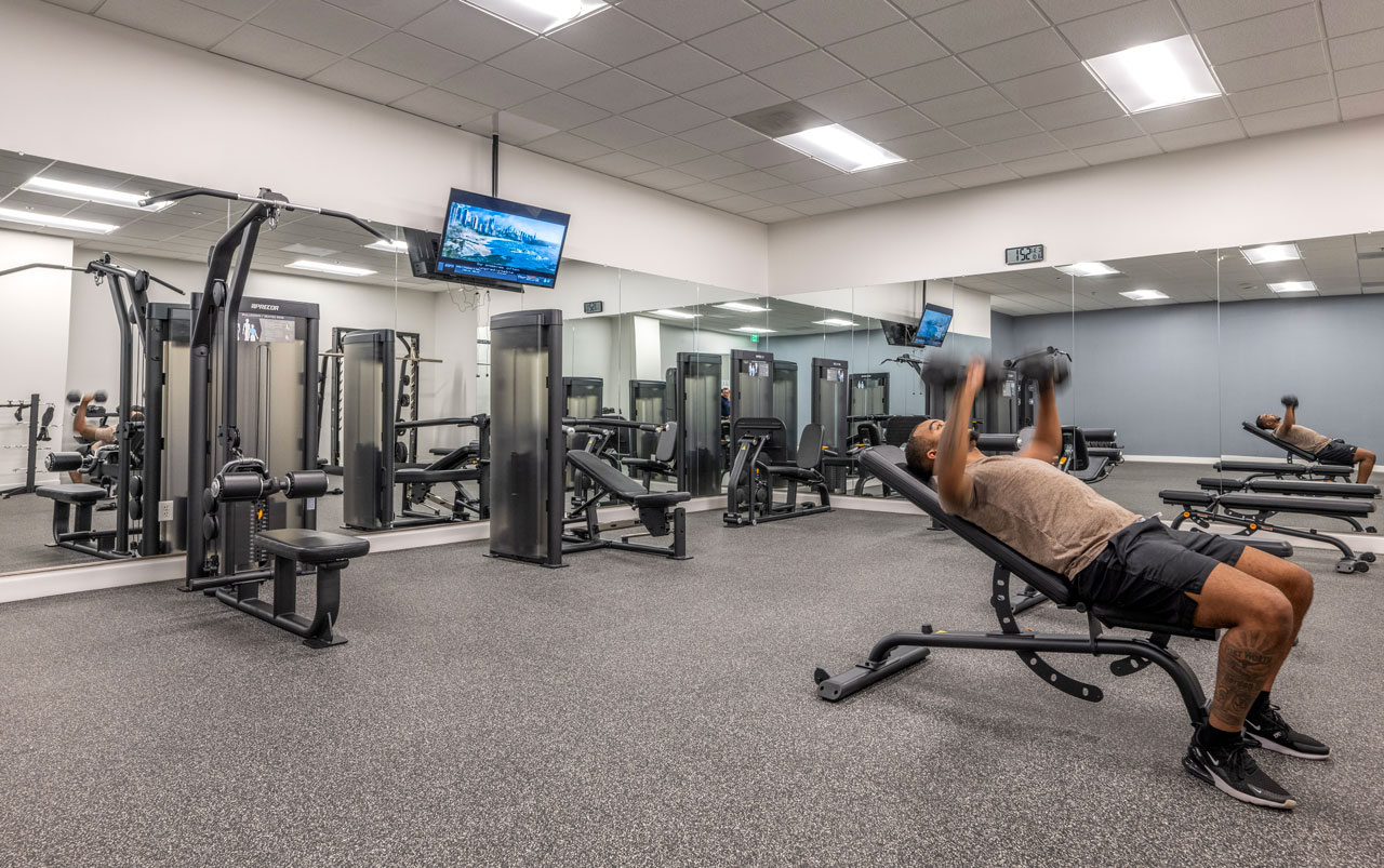 Interior view of the Las Colinas Corporate Center fitness center, featuring modern gym equipment. In the foreground, a person is bench pressing with a barbell on a flat bench. The space is bright and spacious, with clean flooring, mirrors along the walls, and additional strength training machines visible in the background.