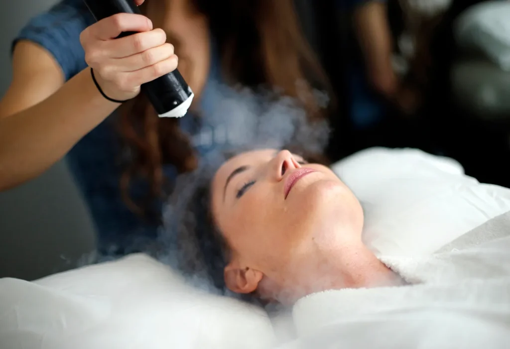 Person receiving localized cryotherapy treatment on their face using a handheld device emitting cold vapor, with visible frost-like mist surrounding the treated area.