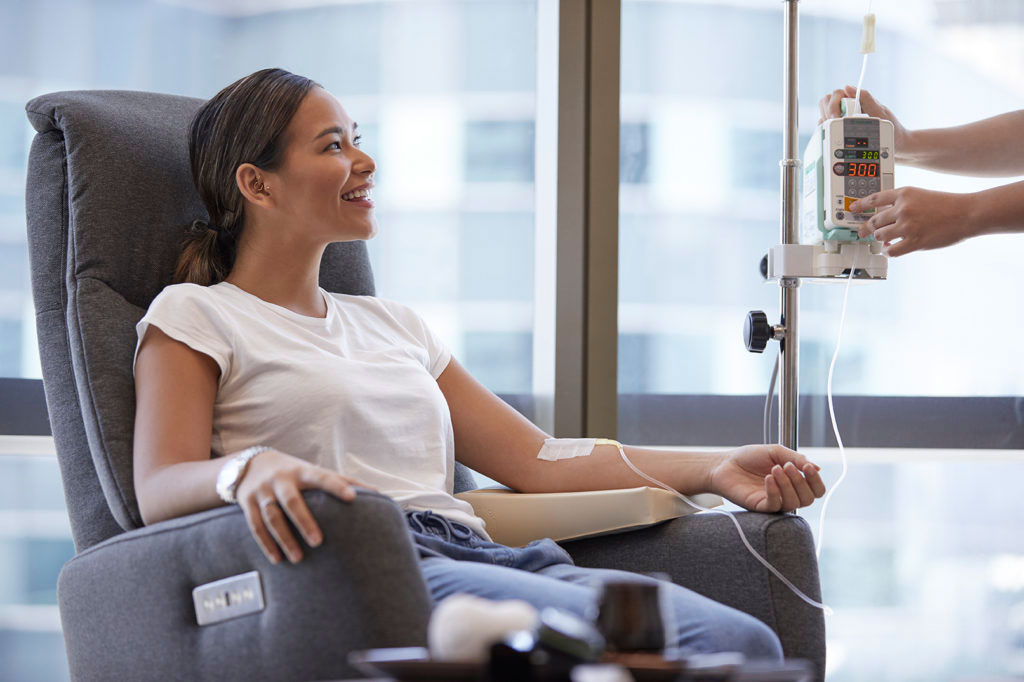 A person sitting in a chair with an IV bag hanging next to them, receiving an intravenous infusion through a needle inserted into their arm.