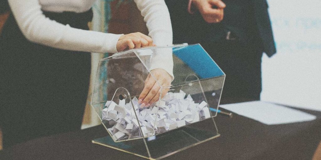 An image of a raffle draw with a glass tumbler filled with raffle tickets. A person is reaching into the bowl to draw a ticket, while excited attendees watch eagerly in the background
