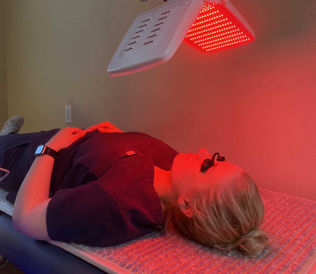 Person lying down on a treatment table with a red light therapy panel positioned above them, casting a warm red glow over their body in a dimly lit room.